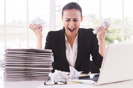 That is too much for me! Furious young woman in formalwear holding papers in hands and shouting while sitting at her working placeの写真素材