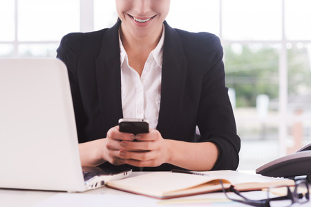 Typing business message. Close-up of young woman in formalwear holding mobile phone while sitting at her working placeの写真素材