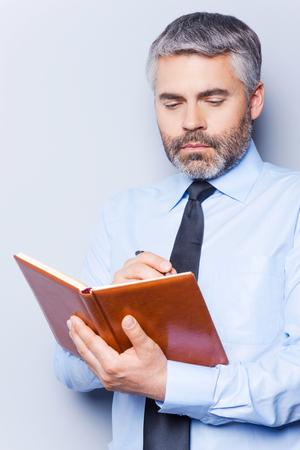 Writing To-Do list. Confident mature man in shirt and tie writing something in note pad while standing against grey backgroundの写真素材