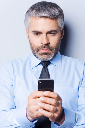Typing business message. Confident mature man in shirt and tie holding mobile phone and looking at it while standing against grey background の写真素材