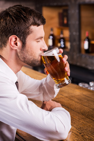 Enjoying his favorite lager. Side view of handsome young man drinking beer while sitting at the bar counter の写真素材