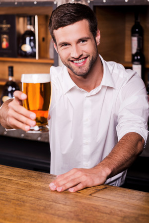 Let me quench your thirst! Handsome young male bartender in white shirt stretching out glass with beer and smiling while standing at the bar counterの写真素材