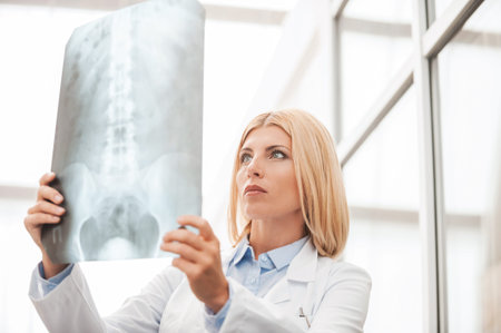 Low angle view of confident female doctor in white uniform holding x-ray image and looking at it