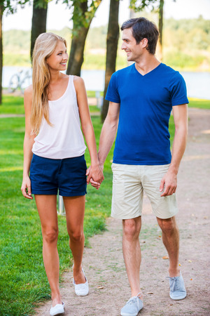 Nice day for date. Full length of beautiful young loving couple walking along the park road and looking at each other with smileの写真素材