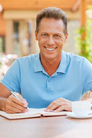 I love working outdoors. Confident mature man writing something in his note pad and smiling while sitting at the table outdoors with house in the background の写真素材