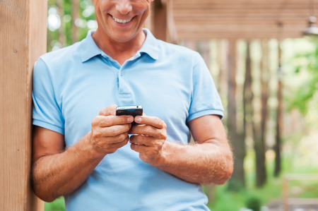 Examining his new smart phone. Cheerful mature man holding mobile phone and smiling while standing outdoors の写真素材