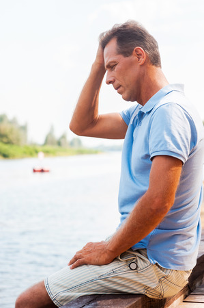 Worried and depressed man. Side view of depressed mature man touching head with hand and looking down while sitting at the quayside の写真素材