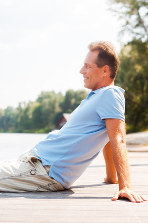 Enjoying doing nothing. Side view of cheerful mature man looking away and smiling while sitting at the quaysideの写真素材