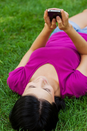 Staying in touch with friends. Attractive young woman typing message on mobile phone and smiling while lying in grass の写真素材