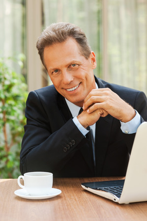 Confident businessman. Confident mature man in formalwear smiling and looking at camera while sitting at the table with laptop on it の写真素材