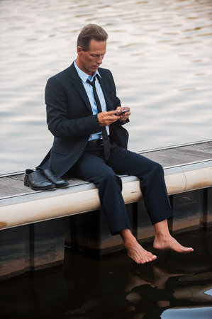 Feeling lonely. Frustrated mature businessman looking at his mobile phone while sitting barefoot at the quayside の写真素材
