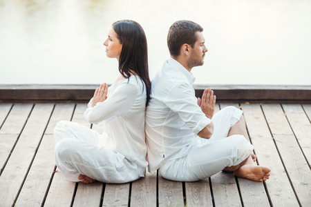 In peace with universe. Side view of beautiful young couple in white clothing meditating outdoors together while sitting back to back in lotus positionの写真素材