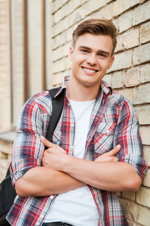 Enjoying time in college. Handsome young man carrying backpack on one shoulder and smiling while leaning at the brick wall の写真素材