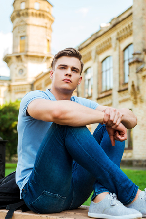 Thinking about future. Thoughtful male student sitting on the bench and looking away with university building in the backgroundの写真素材
