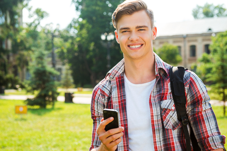 Confident and successful student. Handsome male student holding mobile phone and smiling while standing outdoorsの写真素材