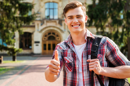 Thumb up for success! Handsome male student showing his thumb up and smiling while standing outdoors with university building in the backgroundの写真素材