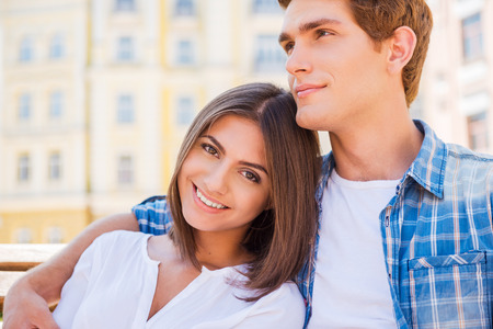 Enjoying their time together. Beautiful young loving couple sitting on the bench together while woman looking at camera and smiling の写真素材