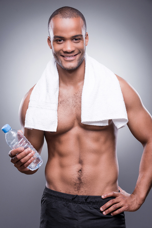 Young shirtless African man holding bottle with water and smiling while standing against grey backgroundの写真素材