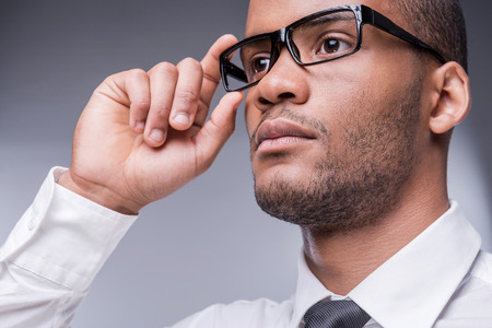 Confident businessman. Low angle view of thoughtful young African man in shirt and tie adjusting his eyeglasses and looking away while standing against grey background の写真素材