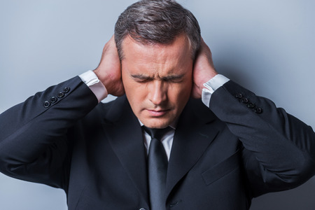 This is too loud! Frustrated mature man in formalwear holding head in hands and keeping eyes closed while standing against grey backgroundの写真素材