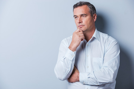 Lost in thoughts. Portrait of thoughtful mature man in shirt holding hand on chin and looking away while standing against grey backgroundの写真素材