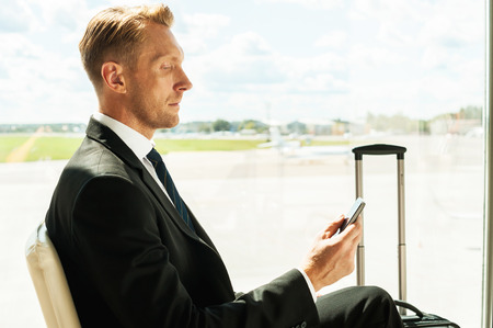 Businessman in airport. Side view of confident businessman in formalwear holding mobile phone while waiting for a flight in airport の写真素材