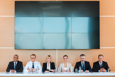 Confident business team. Group of business people sitting together at the table and smiling with large monitor upon their heads の写真素材
