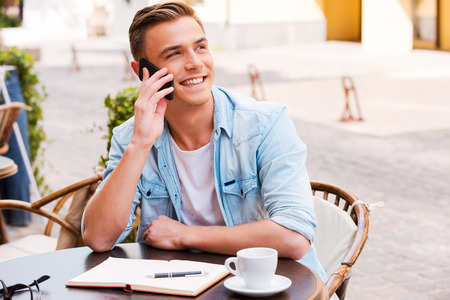 Talking with friends. Confident young man talking on the mobile phone and smiling while sitting in sidewalk cafeの写真素材