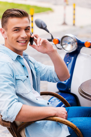 Talking with friend. Relaxed young man talking on the mobile phone and smiling while sitting in sidewalk cafe with scooter in the backgroundの写真素材