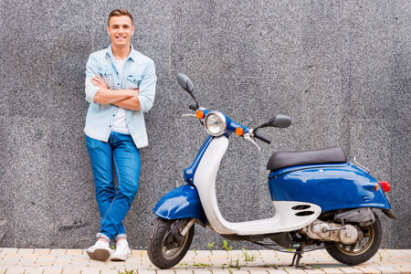 Me and my new scooter. Handsome young man leaning at the wall and smiling while standing near his...