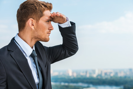 Looking for new business opportunities. Thoughtful young man in formalwear holding hand on forehead and looking away while standing outdoors with cityscape in the backgroundの写真素材