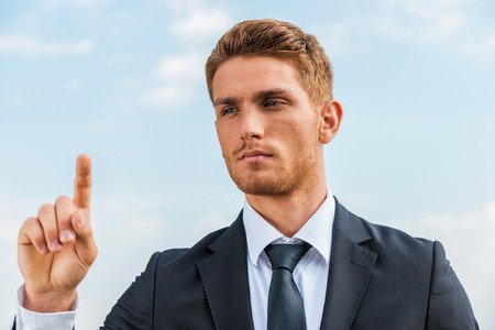 Working on transparent wipe board. Confident young man in formalwear touching a transparent wipe board while standing against sky backgroundの写真素材