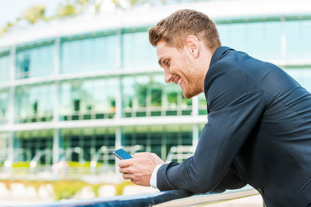 Typing business message. Side view of handsome young man in formalwear holding mobile phone and looking at it with smile while standing outdoorsの写真素材