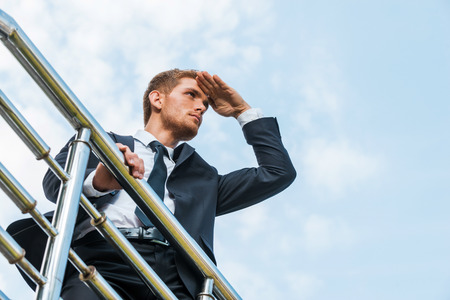 Looking for new opportunities. Low angle view of confident young man in formalwear holding hand on forehead and looking away while standing outdoors and leaning at the metal railingの写真素材