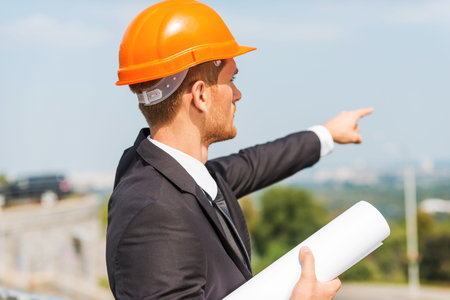 Planning a new construction. Rear view of confident young man in hardhat holding blueprint and...