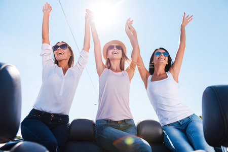 What a great day! Low angle view of three beautiful young women enjoying road trip in convertible...