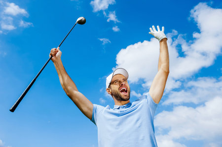 I won! Low angle view of young happy golfer holding driver and raising his arms with blue sky as backgroundの写真素材