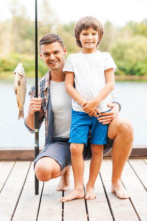 Look what we caught! Father and son looking at camera and smiling while man holding fishing rod with big fish on the hook の写真素材