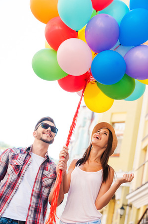 Living a colorful life. Beautiful young loving couple carrying multi colored balloons and smiling while standing outdoors の写真素材