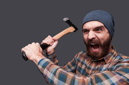 Furious lumberjack. Furious young bearded man swinging his axe and shouting while standing against...