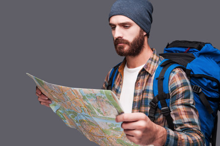 Tourist examining map. Handsome young bearded man carrying backpack and examining map while standing...