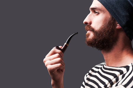 Just me and my pipe. Side view of confident young bearded man in striped clothing holding a smoking pipe and looking away while standing against grey backgroundの写真素材