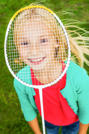 I am ready to play! Top view of cute little girl hiding her face behind badminton racket and smiling while standing on green grassの写真素材