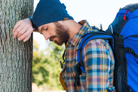 Tired traveler. Side view of tired young man with backpack leaning at the tree and keeping eyes closedの写真素材