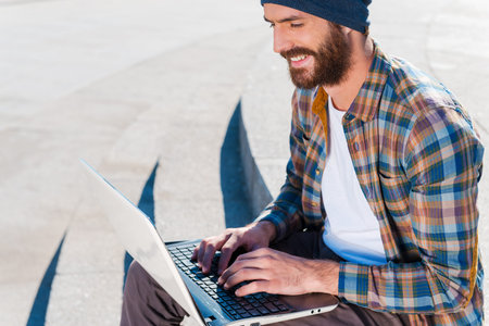 Pleasant working outdoors.  Handsome young bearded man smiling while working on laptop