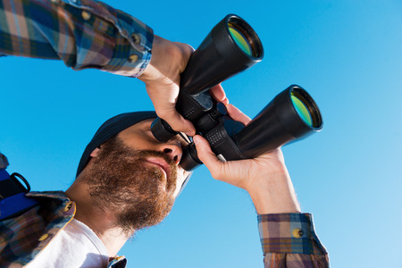 Taking a closer look. Long angle view of handsome young bearded man looking through binoculars awayの写真素材
