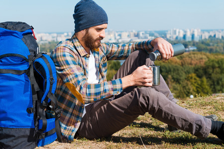 Finally I can relax.  Handsome young man sitting near backpack and pouring tea into a cupの写真素材