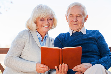 We love reading together. Cheerful senior couple reading a book together while sitting on the park benchの写真素材
