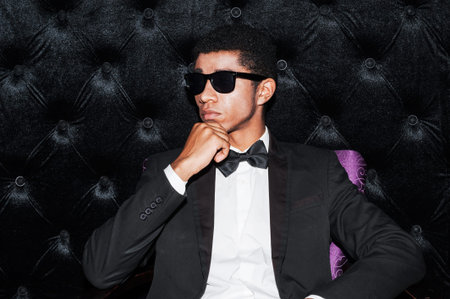 Individual style. Fashionable young Afro-American man sitting on the chair holding hand on chinの写真素材