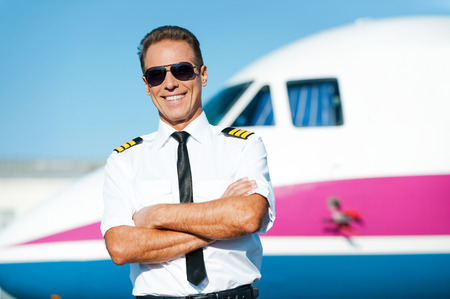 Confident pilot. Confident male pilot in uniform keeping arms crossed and smiling with airplane in the backgroundの写真素材